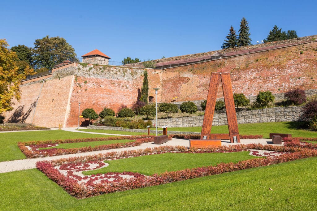 Krajinka Cheb. Udržovaný zahradní záhon s geometrickým vzorem pod hradbami Chebského hradu, trávník, dřevěný rám a kamenné hradby v pozadí.